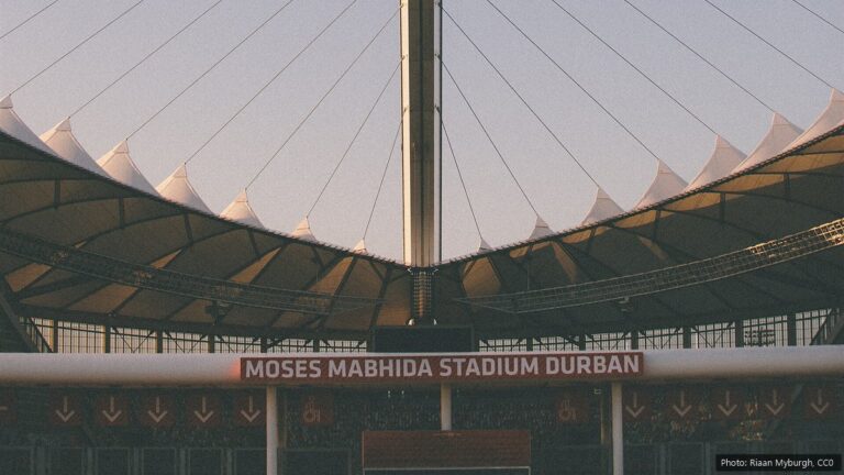 AmaZulu FC: Coach Arthur Zwane, Stadium, Trophies and the 2026/27 Season The white pointed roof and central arch leg of Moses Mabhida Stadium in Durban above a red sign with the stadium's name